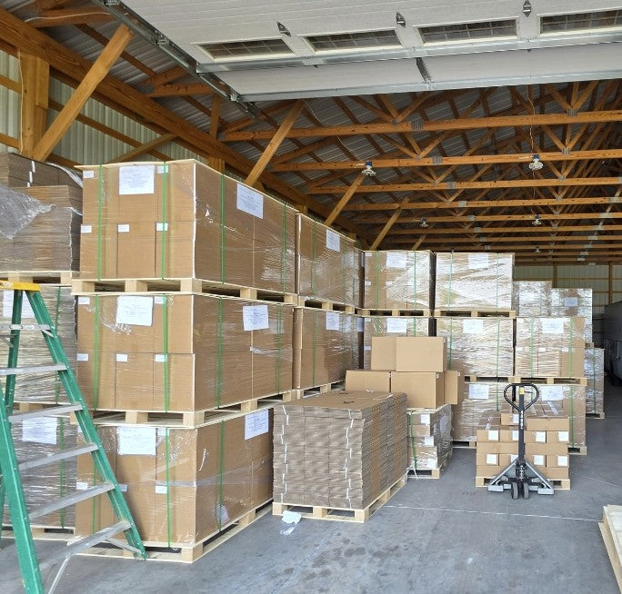 A warehouse interior filled with stacked brown cardboard boxes on wooden pallets, wrapped in clear plastic. A green ladder is visible on the left, a pallet jack stands on the right, and the space has a high ceiling with exposed wooden beams and metal panels.