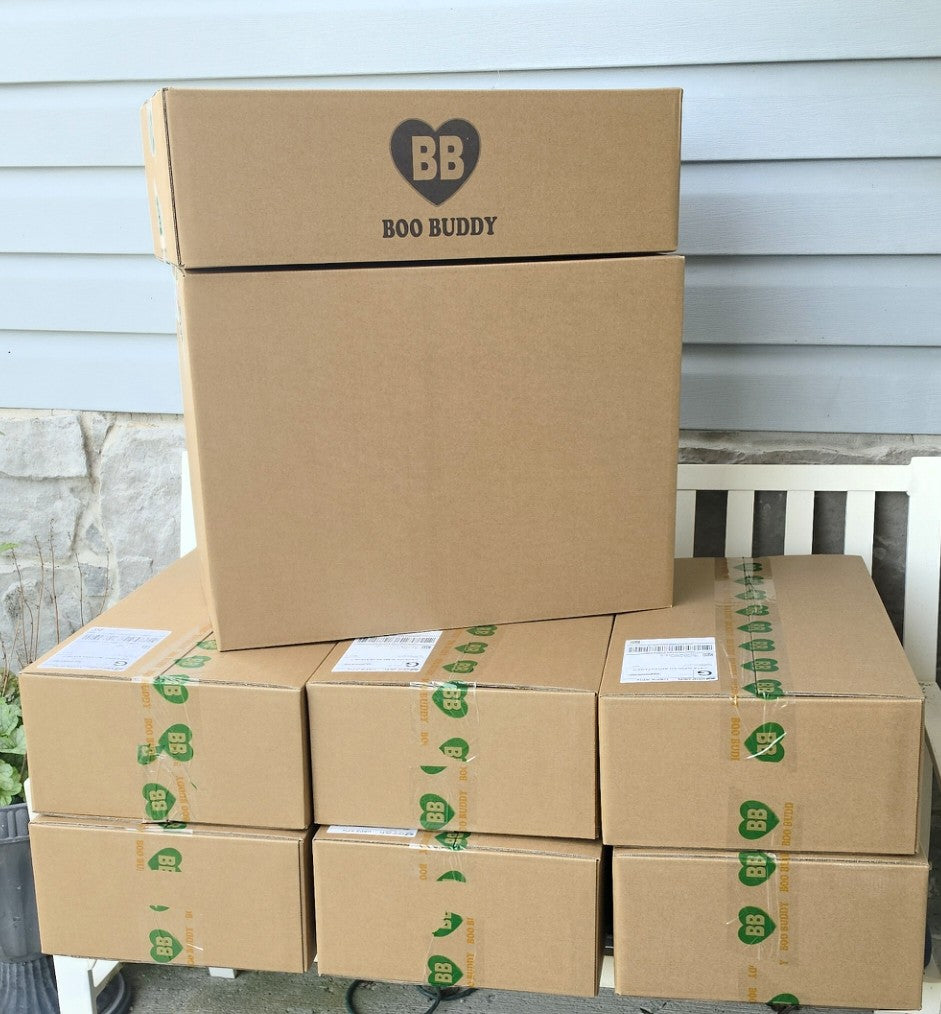 A stack of brown cardboard shipping boxes, with the top box printed with a black heart logo containing 'BB' and the text 'BOO BUDDY'. Several smaller boxes are stacked underneath on a porch or patio area with a light gray siding wall and a white bench visible in the background.