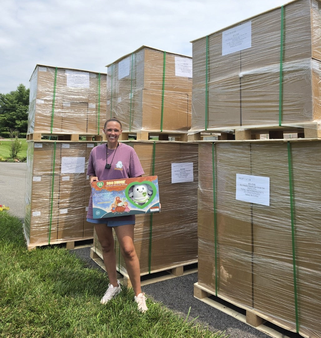 A woman stands outdoors on grass beside large wrapped pallet loads of goods, holding a colorful toy box. The scene includes grass, pavement, trees, and stacked shipping pallets under a cloudy sky.