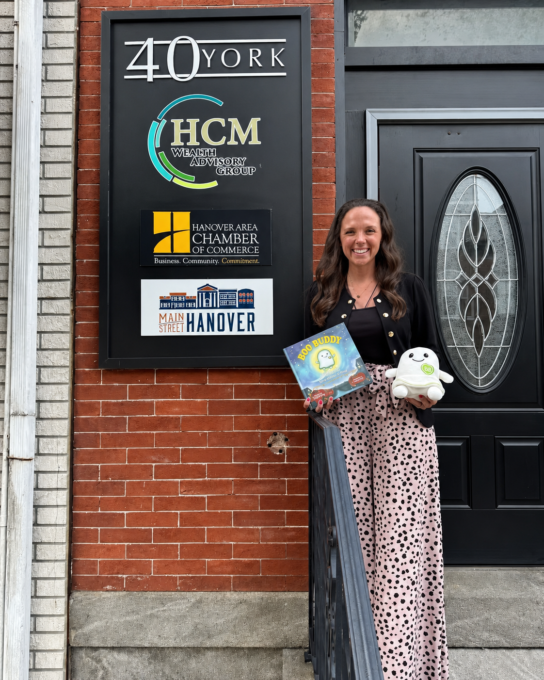 A woman stands in front of a brick building door holding a book and a plush toy. A large black sign on the wall reads 40 York, HCM, and includes other business names including Danceur Area Chamber of Commerce and Main Hanover.