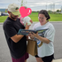 A man and a woman outdoors holding a book and a brown paper shopping bag, with a pink heart graphic overlay covering the man's face. The man wears a black t-shirt, maroon shorts, and a beige cap, while the woman wears a white graphic t-shirt and black shorts.