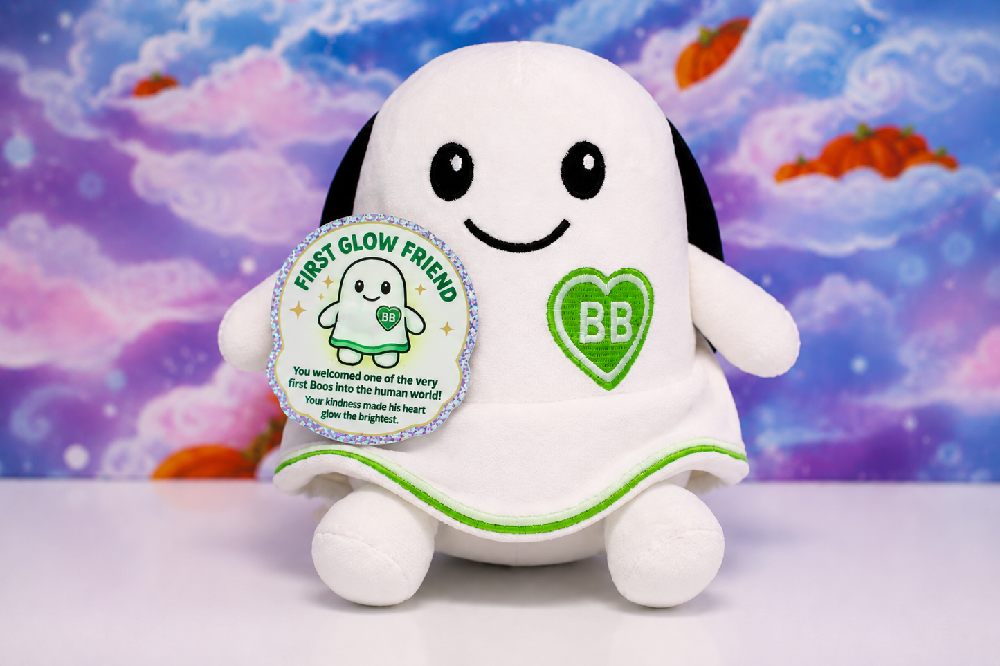 A white plush toy shaped like a smiling ghost, a green heart patch with 'BB' on the chest, and a round silver-bordered tag reading 'FIRST GLOW FRIEND,' set against a colorful cloud-like backdrop.