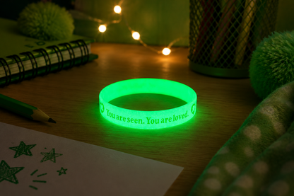 A glowing green silicone wristband with the printed message 'You are seen. You are loved.